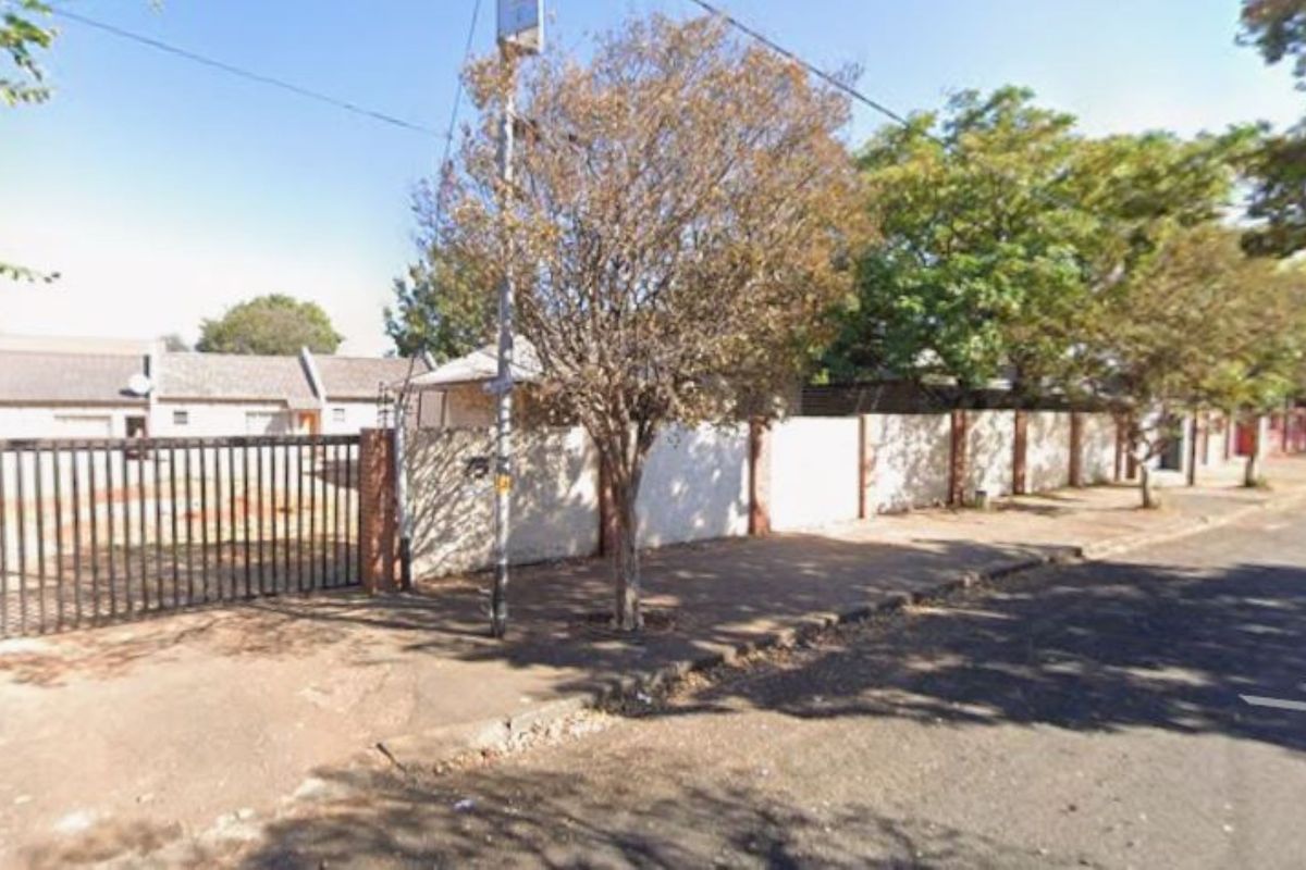 Street view showing the property perimeter wall, metal entrance gate, and adjacent sidewalk area.
