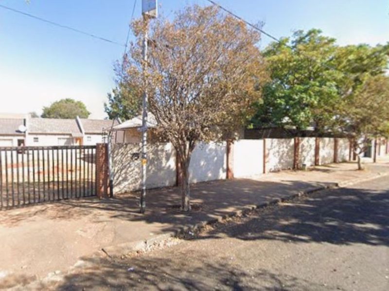 Street view showing the property perimeter wall, metal entrance gate, and adjacent sidewalk area.