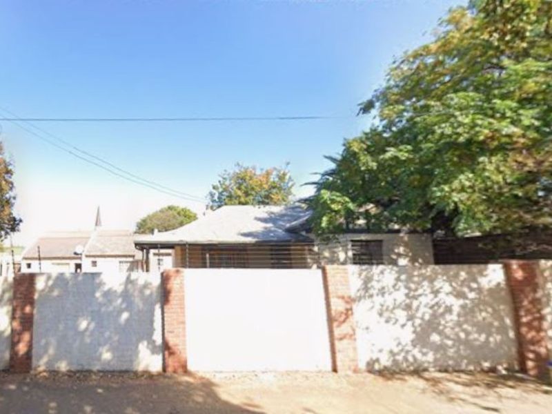 Front view of the property featuring a solid perimeter wall with brick pillars and security fencing, partially obscured by mature trees.