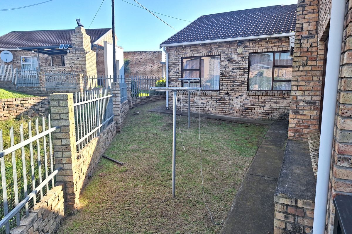 A rear exterior view of a face brick house featuring a small lawn area, concrete pathway, and metal perimeter fencing.