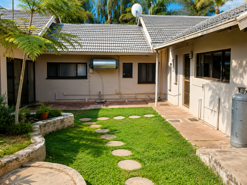 Rear courtyard area featuring a lawn, stone retaining wall, paved pathways, and external utility installations including a geyser and gas cylinder.