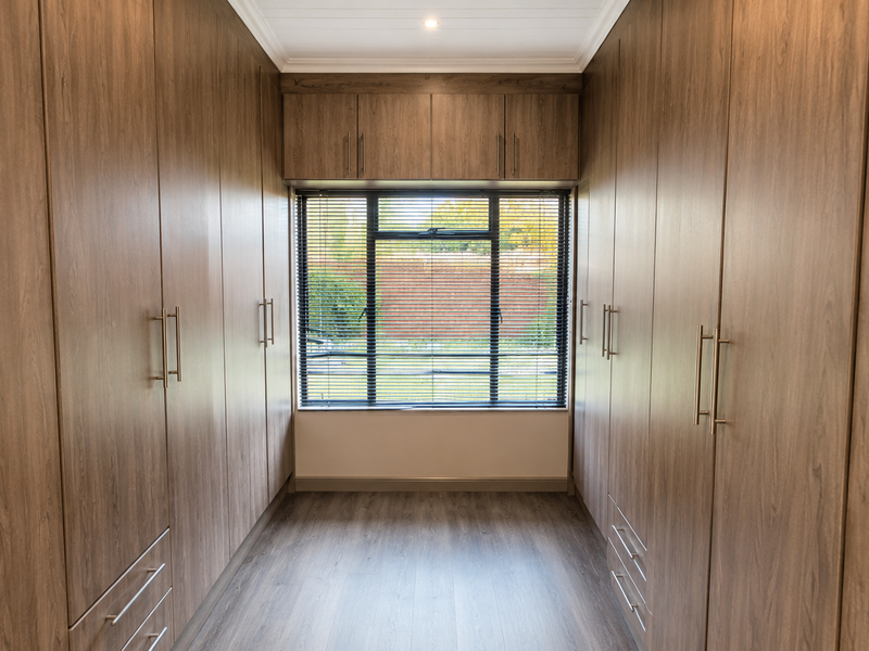 A well-appointed walk-in dressing room featuring extensive floor-to-ceiling built-in cabinetry, modern laminate flooring, and a window with fitted venetian blinds.