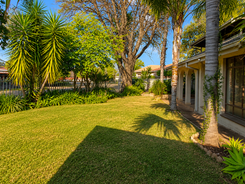 A well-maintained garden featuring a lush lawn, mature trees, and a paved walkway leading to a covered patio area.