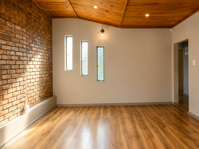 A bright, empty living room featuring a striking exposed brick feature wall, warm timber ceiling, and modern laminate flooring with unique vertical windows.