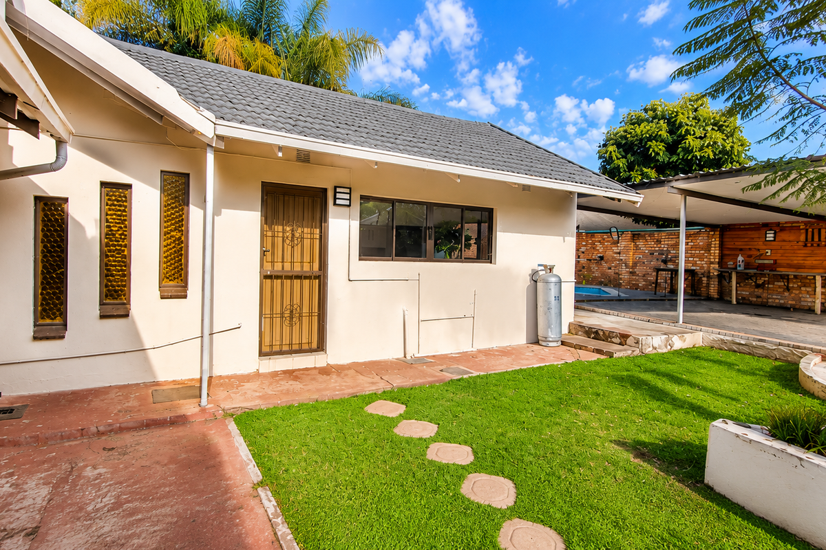 Rear exterior view featuring a paved patio, lawn, raised garden beds, and a covered carport area with brick boundary walls.