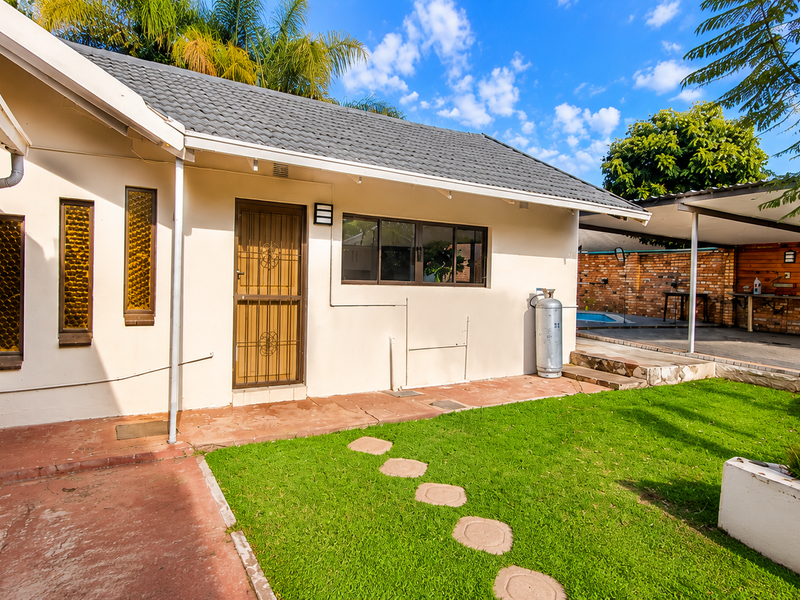 Rear exterior view featuring a paved patio, lawn, raised garden beds, and a covered carport area with brick boundary walls.