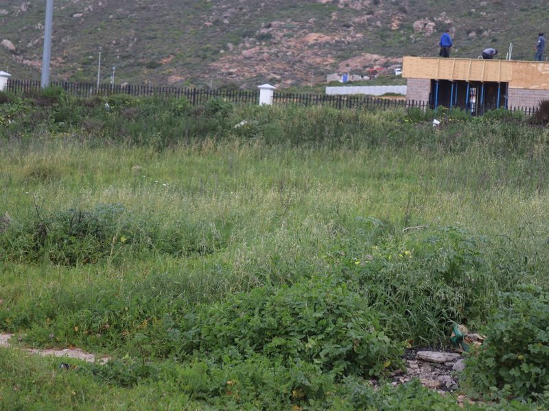 A view of an undeveloped residential plot featuring overgrown grass and perimeter fencing, with neighboring construction visible in the background.