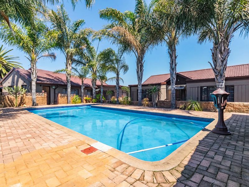 A well-maintained outdoor swimming pool area featuring brick paving, surrounding palm trees, and a secure boundary wall.