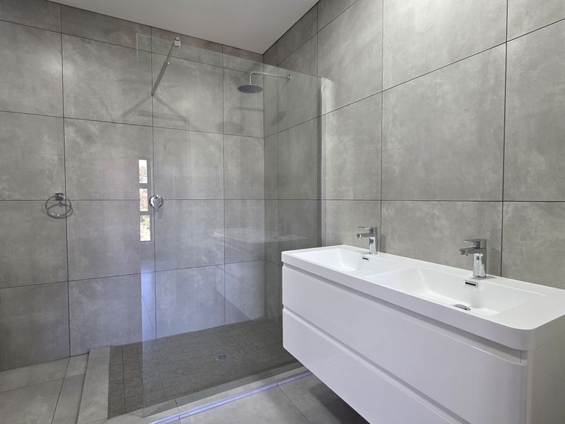 Modern bathroom featuring a double vanity, walk-in shower with glass screen, rain shower head, and contemporary large-format grey tiling throughout.