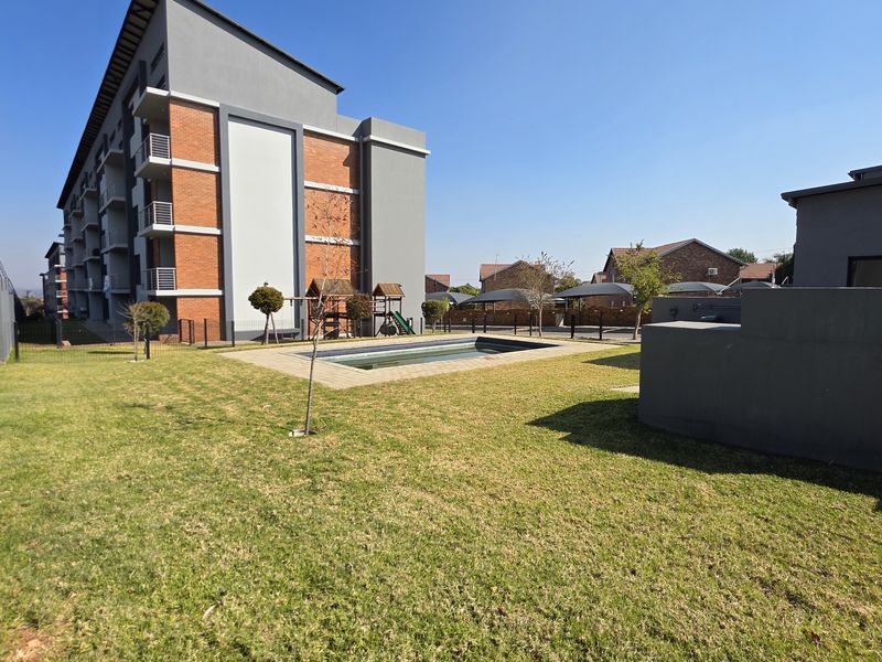 Communal outdoor area featuring a swimming pool, well-maintained lawn, and perimeter security fencing within a modern apartment complex.