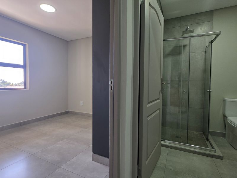 A bright, modern bedroom featuring neutral tiled flooring, recessed lighting, and an adjoining en-suite bathroom with a glass-enclosed shower and contemporary fixtures.