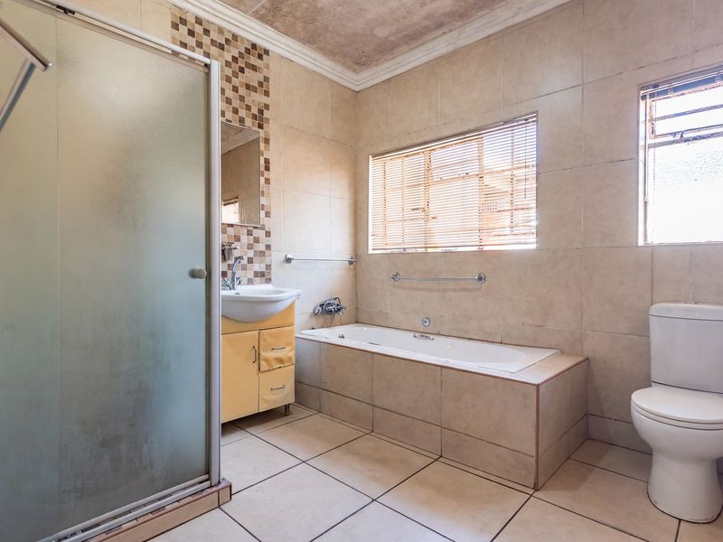 A functional bathroom featuring a built-in bathtub, separate shower enclosure, vanity basin, and toilet, with neutral wall and floor tiling throughout.