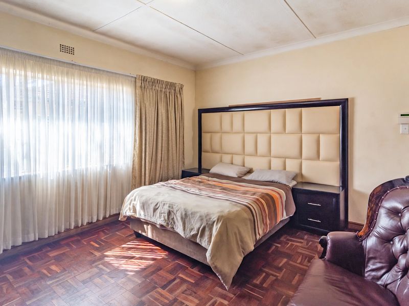 A bedroom featuring classic parquet flooring, a large built-in headboard with integrated bedside tables, and natural light through large windows.