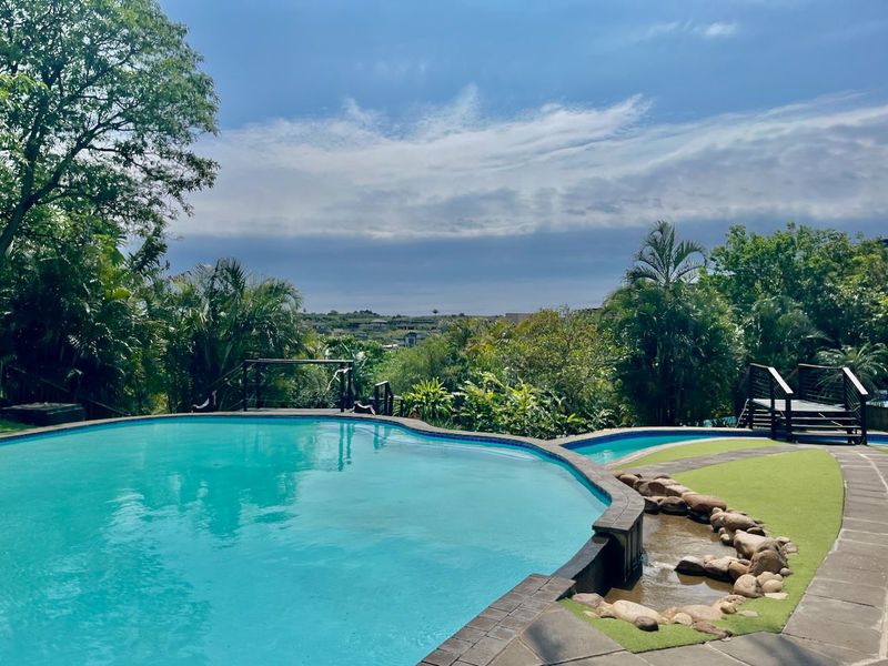 A well-maintained swimming pool area featuring stone landscaping, artificial turf, and elevated decking with scenic views.