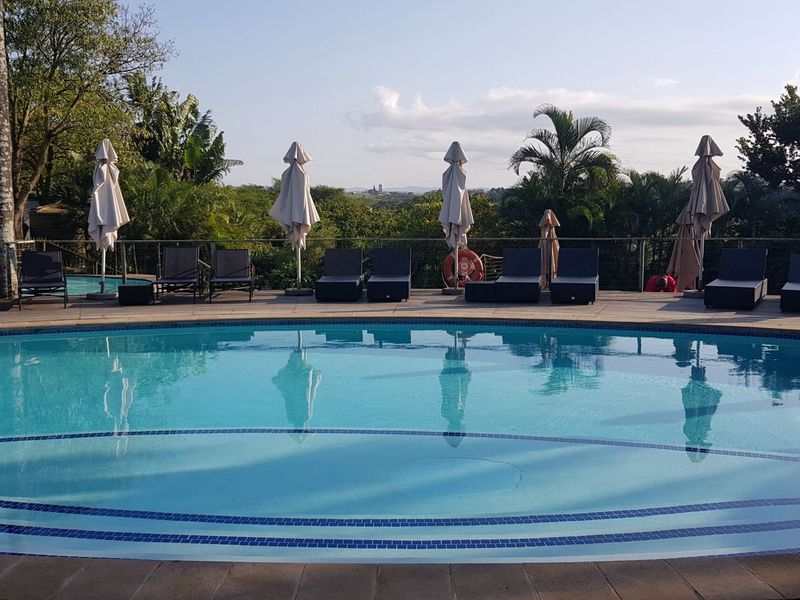 A well-maintained outdoor swimming pool area featuring paved decking, safety railings, and lush surrounding garden views.