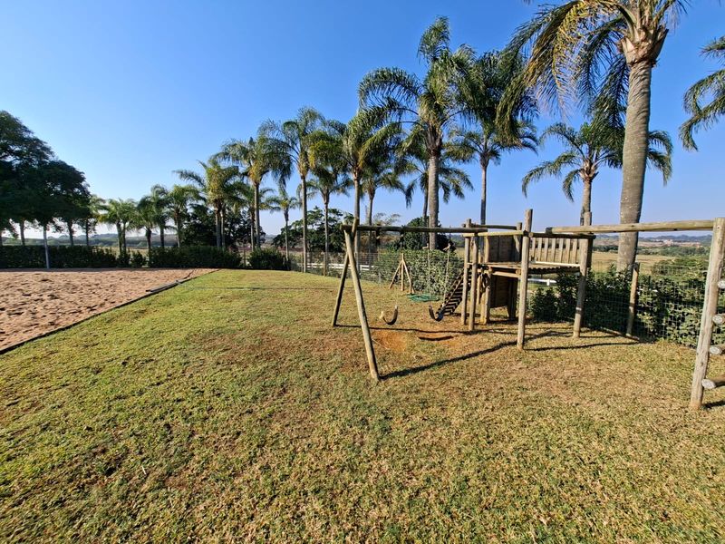 A spacious outdoor garden area featuring a sand arena, mature palm trees, and a wooden playground structure, set against a scenic landscape.