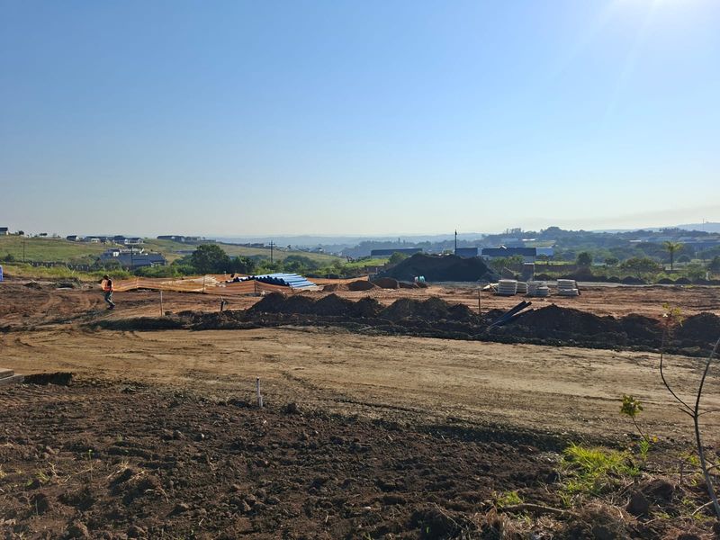 A view of a residential development site showing ongoing earthworks, cleared land, and infrastructure piping in a suburban setting.