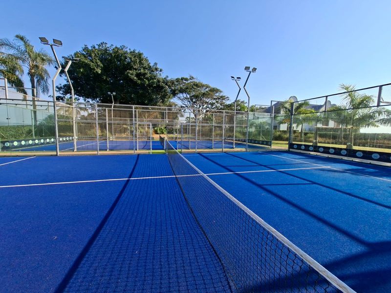 A well-maintained outdoor padel court featuring blue synthetic turf, wire mesh perimeter fencing, and integrated floodlighting for evening play.