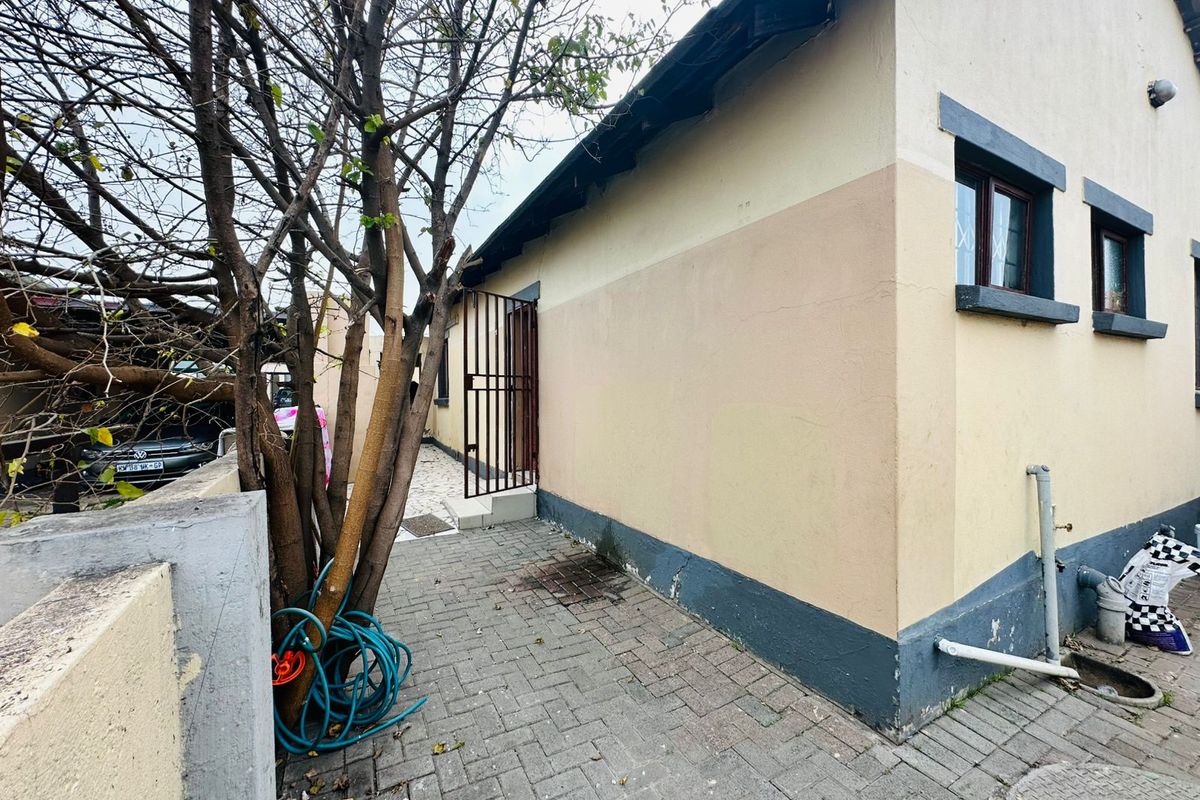 A side view of the property showing a paved walkway, security gate, and exterior wall finishes requiring maintenance.
