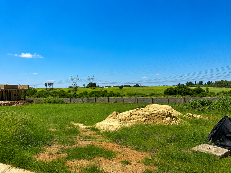 A vacant residential plot featuring a perimeter wall, grassy terrain, and distant power lines under a clear blue sky.