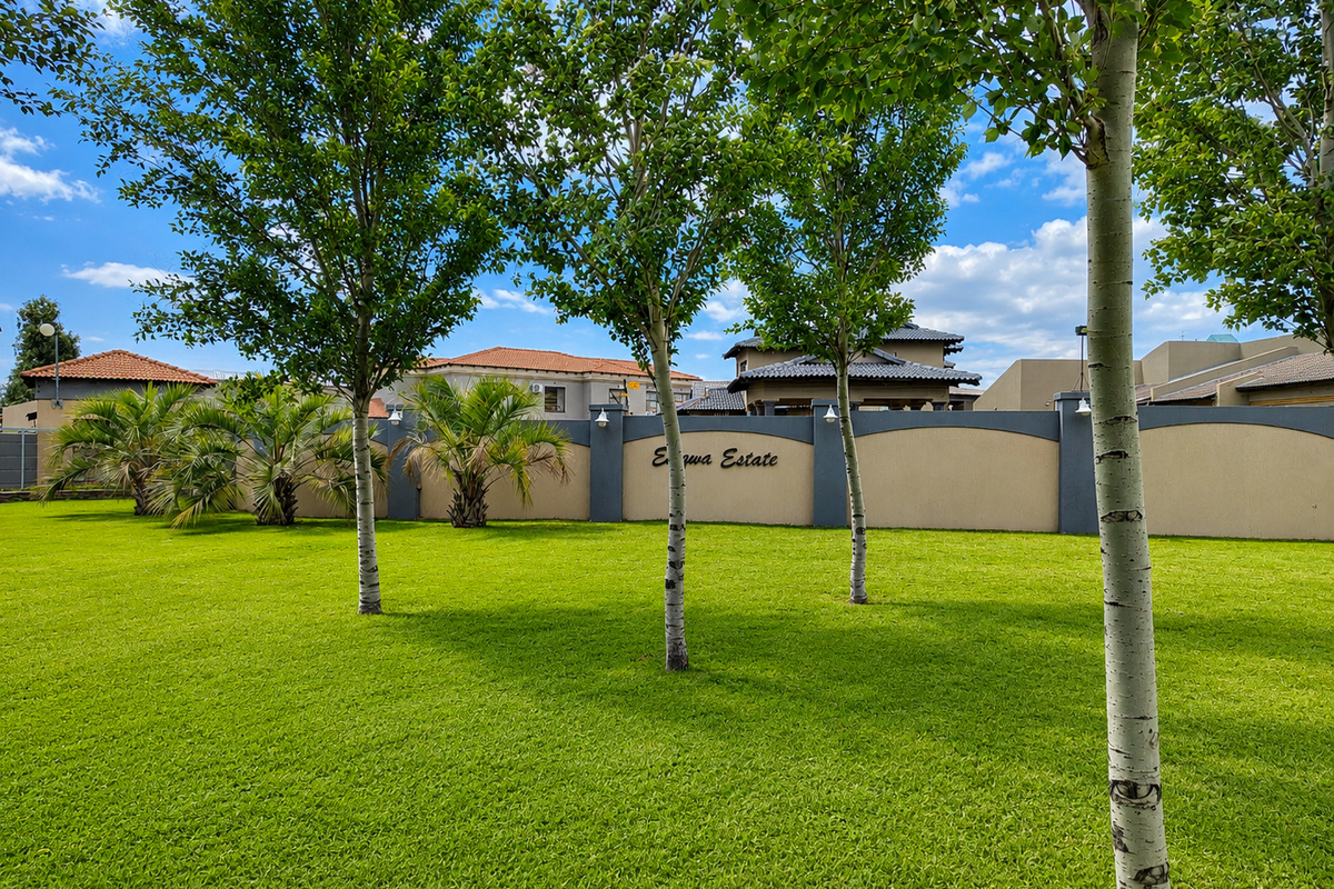 A well-maintained landscaped garden area featuring a manicured lawn, mature trees, and a perimeter boundary wall with estate signage.