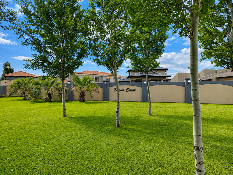 A well-maintained landscaped garden area featuring a manicured lawn, mature trees, and a perimeter boundary wall with estate signage.