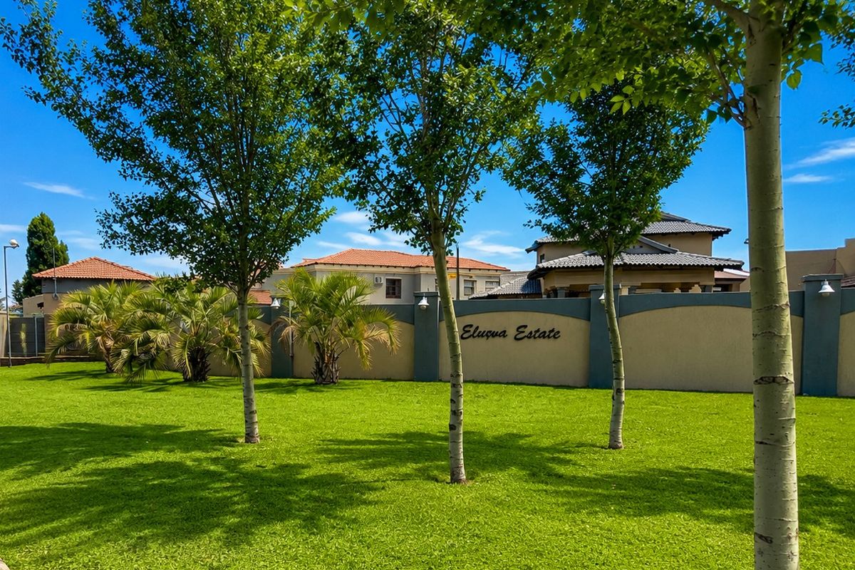 A well-maintained communal garden area featuring lush green lawns, mature trees, and a secure perimeter wall within the Elusua Estate.