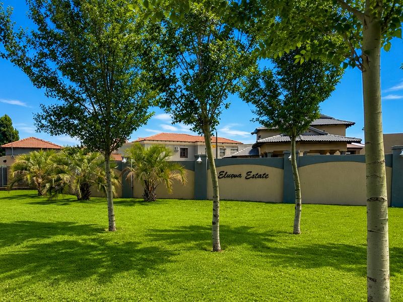 A well-maintained communal garden area featuring lush green lawns, mature trees, and a secure perimeter wall within the Elusua Estate.