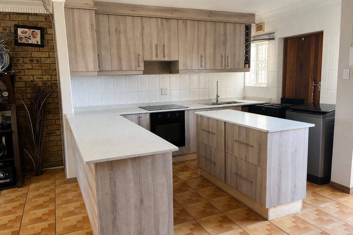 Modern kitchen featuring light wood-grain cabinetry, stone countertops, a central island, and an integrated electric hob and oven, set against a rustic brick feature wall.