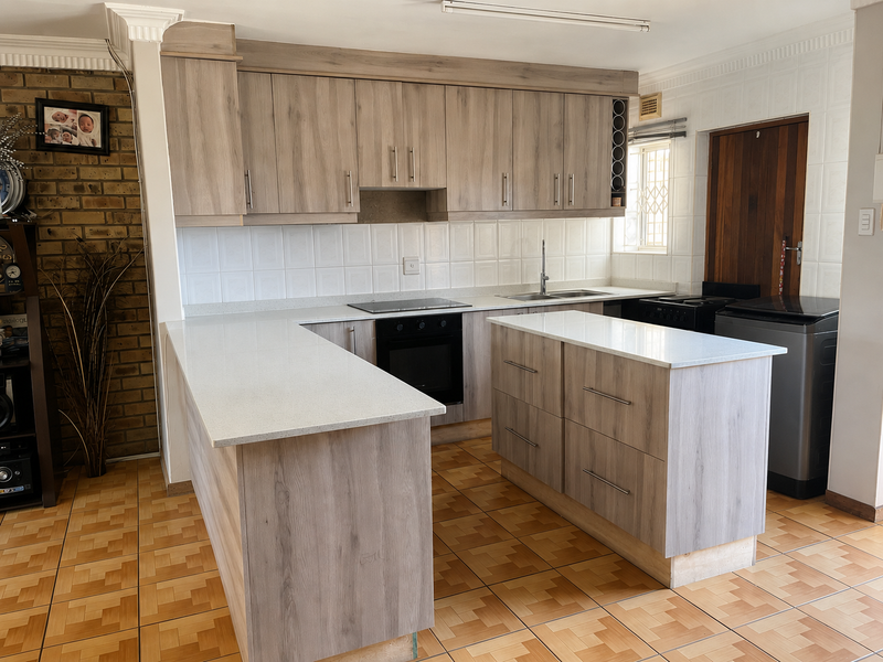 Modern kitchen featuring light wood-grain cabinetry, stone countertops, a central island, and an integrated electric hob and oven, set against a rustic brick feature wall.