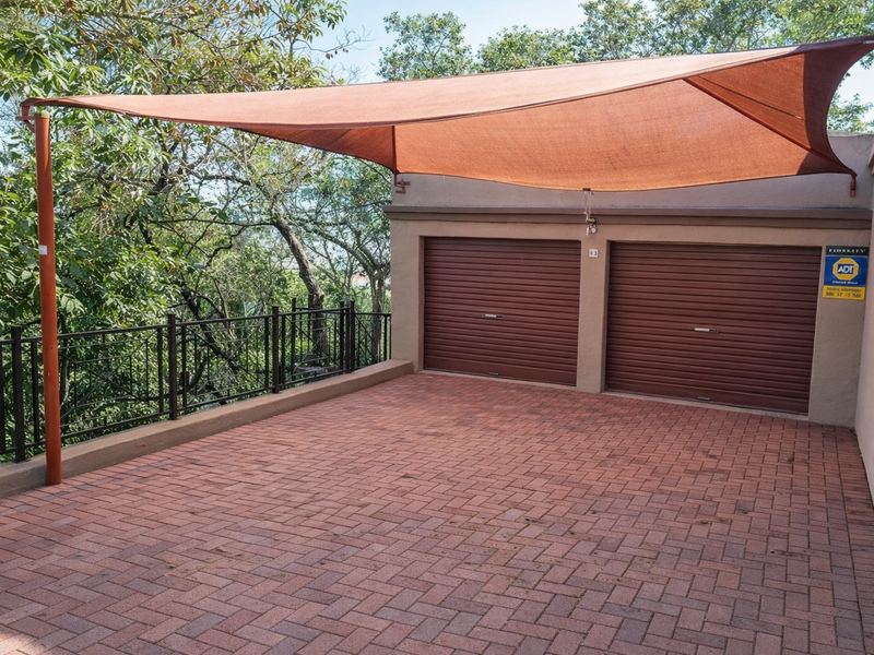 A double garage with roller doors and a paved driveway area featuring a protective shade sail and metal balustrade overlooking the surrounding trees.