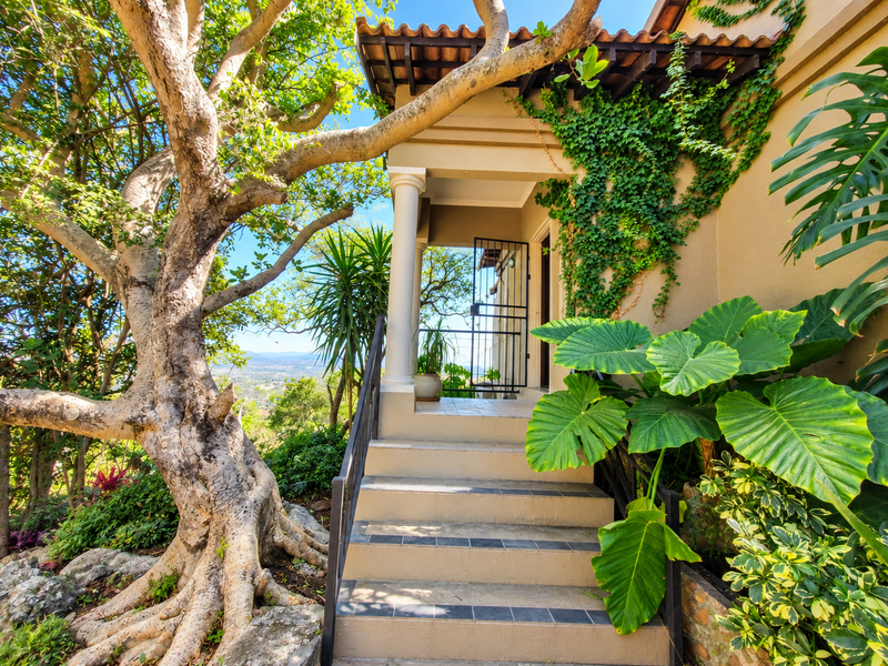 A charming covered entrance porch featuring tiled steps, a metal security gate, and lush surrounding garden foliage with scenic views.