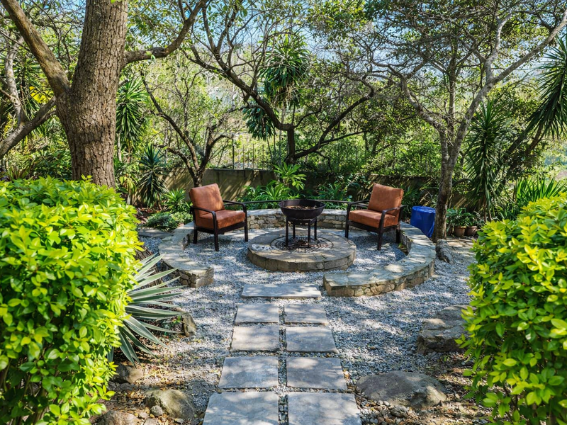 A tranquil garden area featuring a stone-paved path leading to a circular stone fire pit, surrounded by mature trees and lush landscaping.
