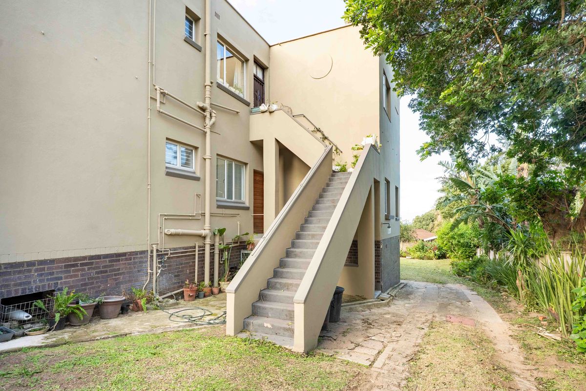 Rear exterior view of the townhouse featuring an external staircase, concrete paving, and established garden surroundings.