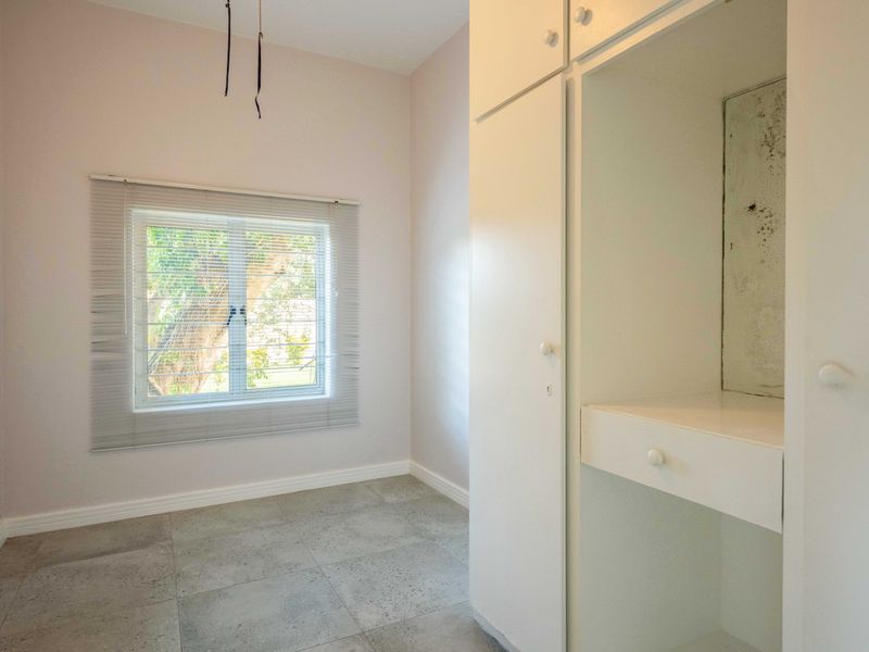 An empty bedroom featuring tiled flooring, a window with fitted blinds, and a built-in wardrobe with internal storage shelving.