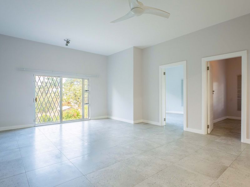 A bright, spacious, and empty living area featuring neutral tiled flooring, a ceiling fan, and a sliding glass door with security gate access.