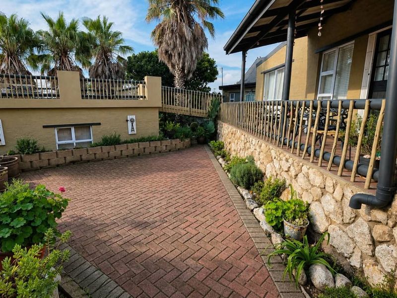 A well-maintained rear exterior area featuring a paved patio, stone retaining wall, and a covered veranda with railing.