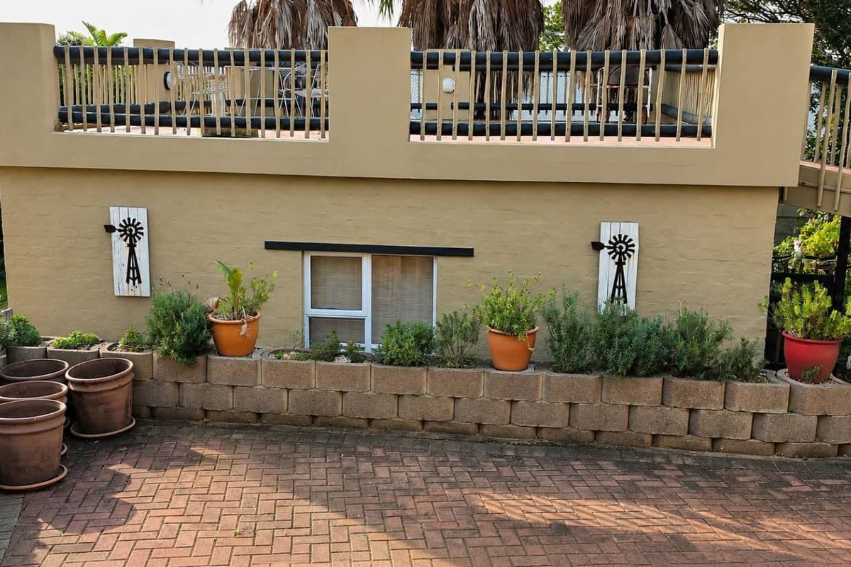 Rear exterior view featuring a paved driveway, a low retaining wall with garden beds, and an elevated balcony with a railing.