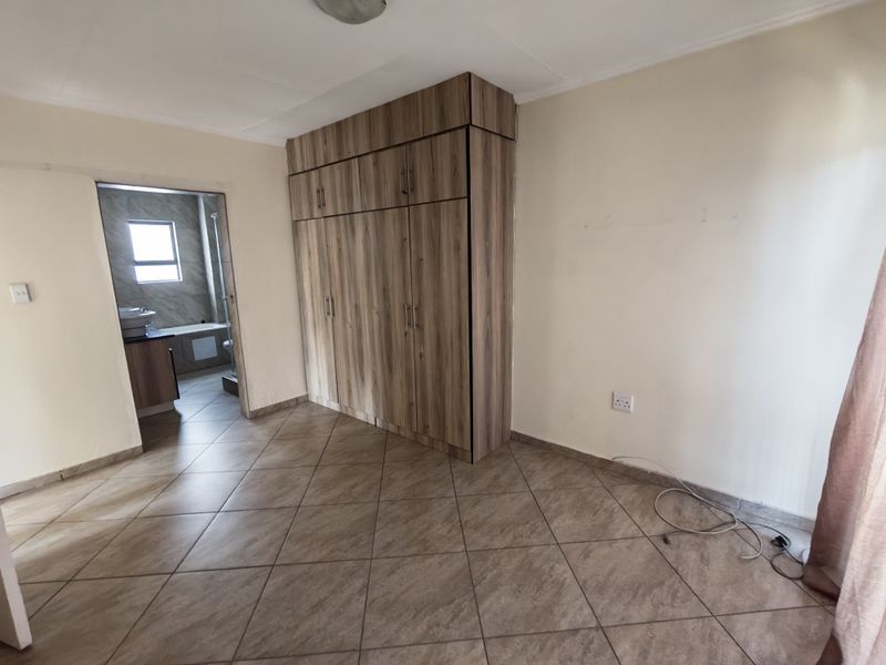 A spacious bedroom featuring neutral tiled flooring and extensive built-in wooden cupboards, with an adjoining en-suite bathroom visible.