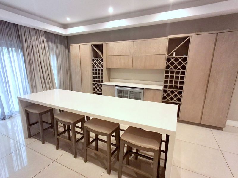 A modern built-in bar area featuring light wood cabinetry, integrated wine storage, an undercounter beverage fridge, and a sleek stone-topped island.