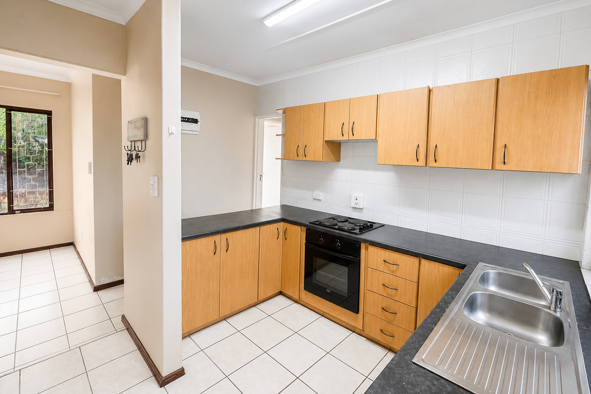 A functional kitchen featuring light wood-tone cabinetry, dark laminate countertops, built-in oven, electric hob, and a double stainless steel sink with tiled flooring.