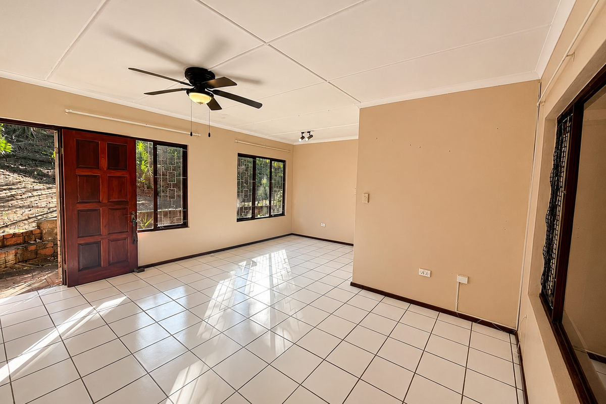 A bright, empty living room featuring white tiled flooring, a ceiling fan, and large windows with security bars, offering direct access to the exterior.