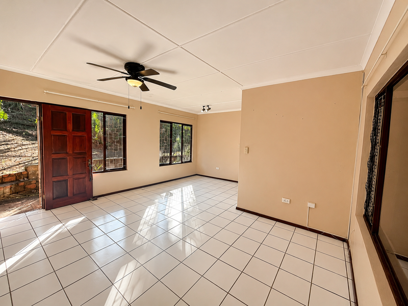 A bright, empty living room featuring white tiled flooring, a ceiling fan, and large windows with security bars, offering direct access to the exterior.
