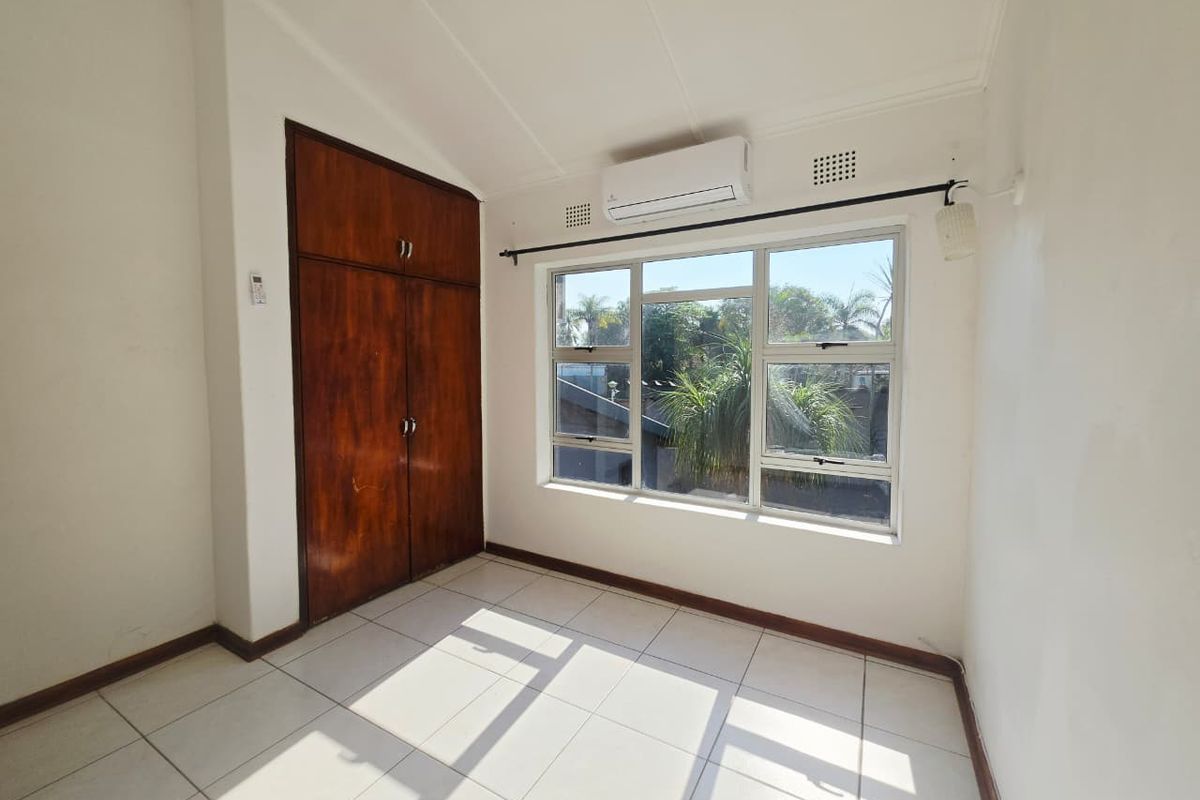 A bright, empty bedroom featuring tiled flooring, a built-in wooden wardrobe, and a wall-mounted air conditioning unit.