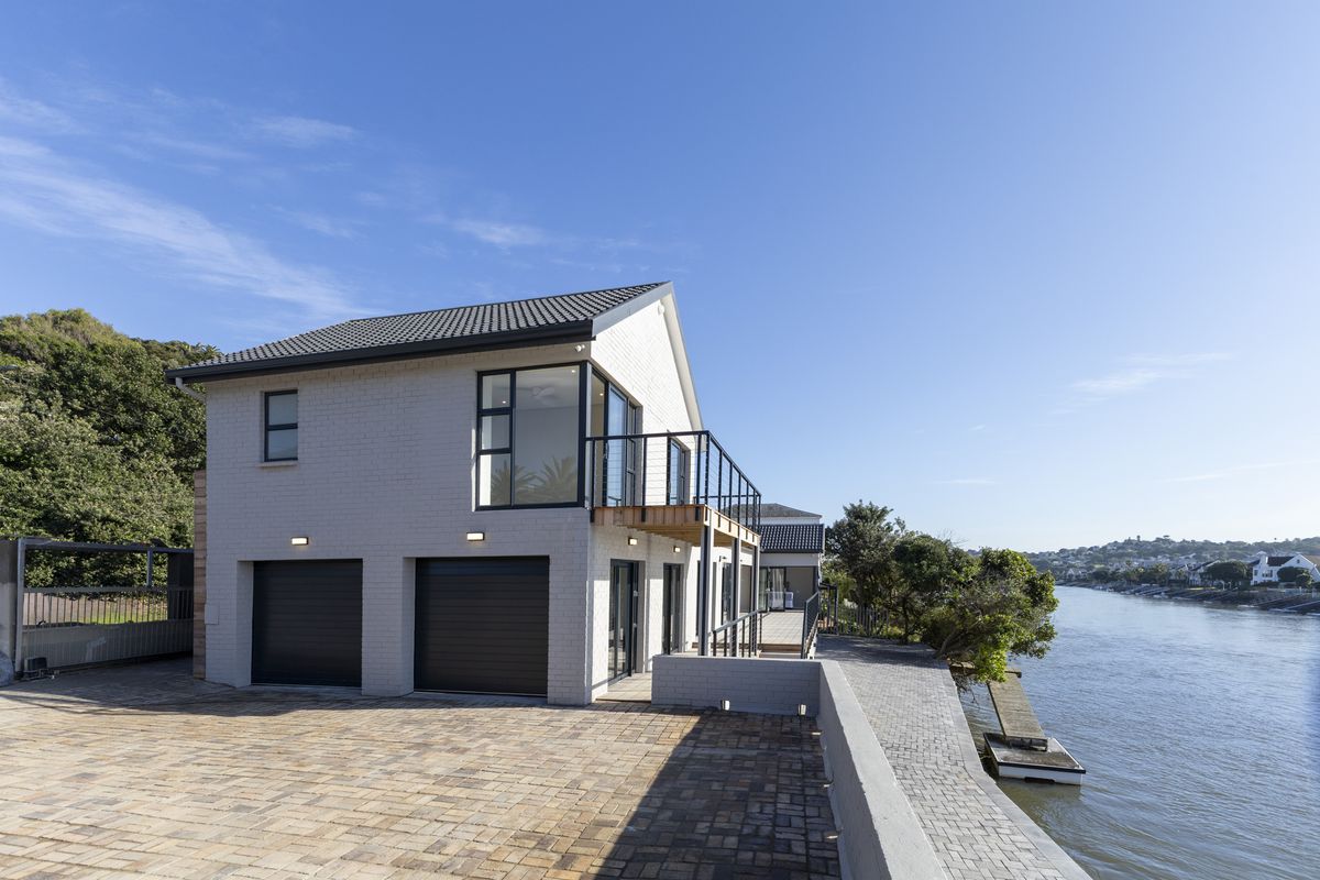 Modern double-storey waterfront home featuring a double garage, upper-level balcony, and paved driveway with direct access to the river.