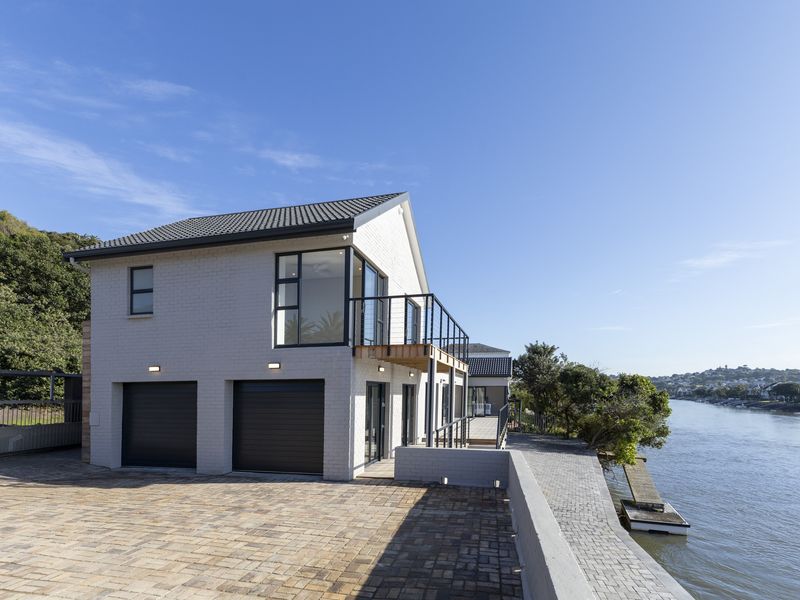 Modern double-storey waterfront home featuring a double garage, upper-level balcony, and paved driveway with direct access to the river.
