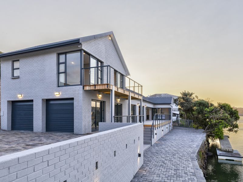 Modern waterfront residence featuring a double garage, elevated balcony, paved outdoor areas, and a private jetty overlooking the water.