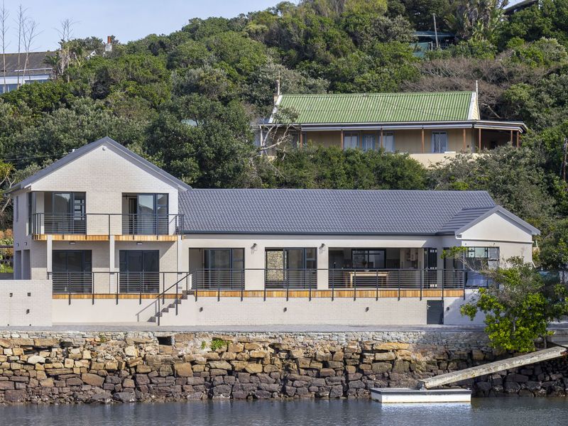 A modern waterfront residence featuring a stone retaining wall, expansive balconies with metal railings, and a private jetty overlooking the water.