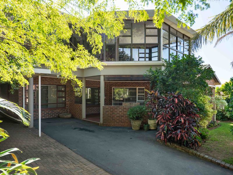 Front view of a multi-level brick house featuring a paved driveway, covered carport, and large windows with security bars, surrounded by established garden greenery.