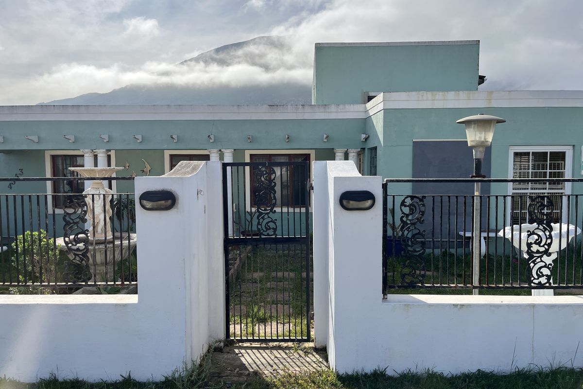 Front elevation of a house featuring a gated entrance, masonry boundary wall, and decorative fountain with mountain views in the background.